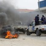 Uma foto de rua capturada durante protestos violentos em Luanda, Angola, mostra um ambiente caótico com fumaça preta espessa preenchendo o ar a partir de pneus queimados no chão. Um veículo de patrulha da polícia de Angola, um pickup truck branco e azul com as palavras "POLÍCIA" e emblemas oficiais visíveis na parte traseira, move-se pela estrada cheia de detritos. Vários policiais em uniformes azuis estão visíveis no veículo e perto dele. Ao fundo, edifícios comerciais e transeuntes podem ser vistos sob um céu nublado. A imagem captura a tensão e o descontentamento social decorrentes do aumento do custo de vida e outras questões socioeconômicas no país.