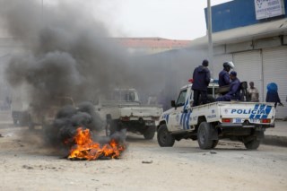 Uma foto de rua capturada durante protestos violentos em Luanda, Angola, mostra um ambiente caótico com fumaça preta espessa preenchendo o ar a partir de pneus queimados no chão. Um veículo de patrulha da polícia de Angola, um pickup truck branco e azul com as palavras "POLÍCIA" e emblemas oficiais visíveis na parte traseira, move-se pela estrada cheia de detritos. Vários policiais em uniformes azuis estão visíveis no veículo e perto dele. Ao fundo, edifícios comerciais e transeuntes podem ser vistos sob um céu nublado. A imagem captura a tensão e o descontentamento social decorrentes do aumento do custo de vida e outras questões socioeconômicas no país.